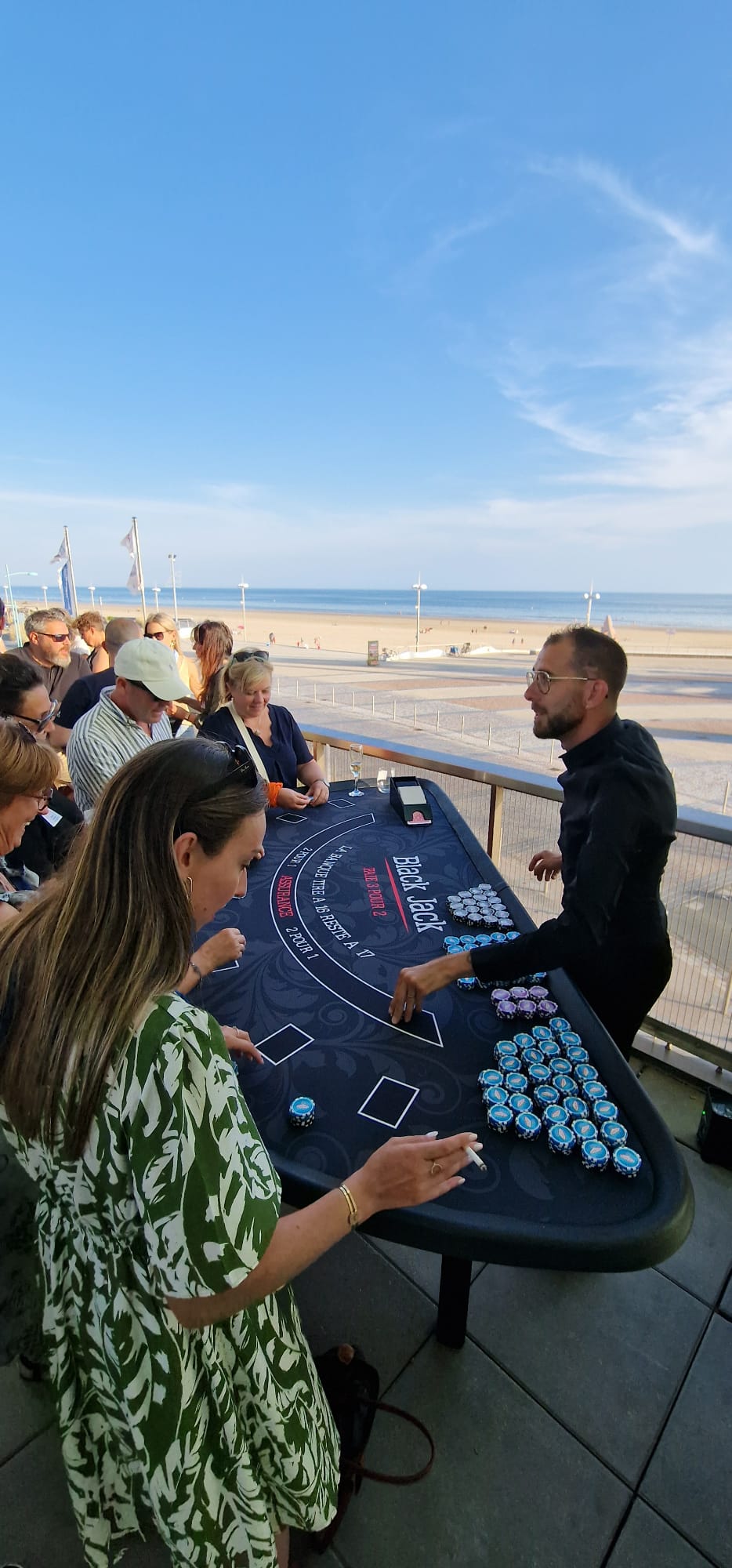 Croupier professionnel animant une table de Black Jack pour un groupe d'invités sur une terrasse en bord de mer lors d'une soirée événementielle.