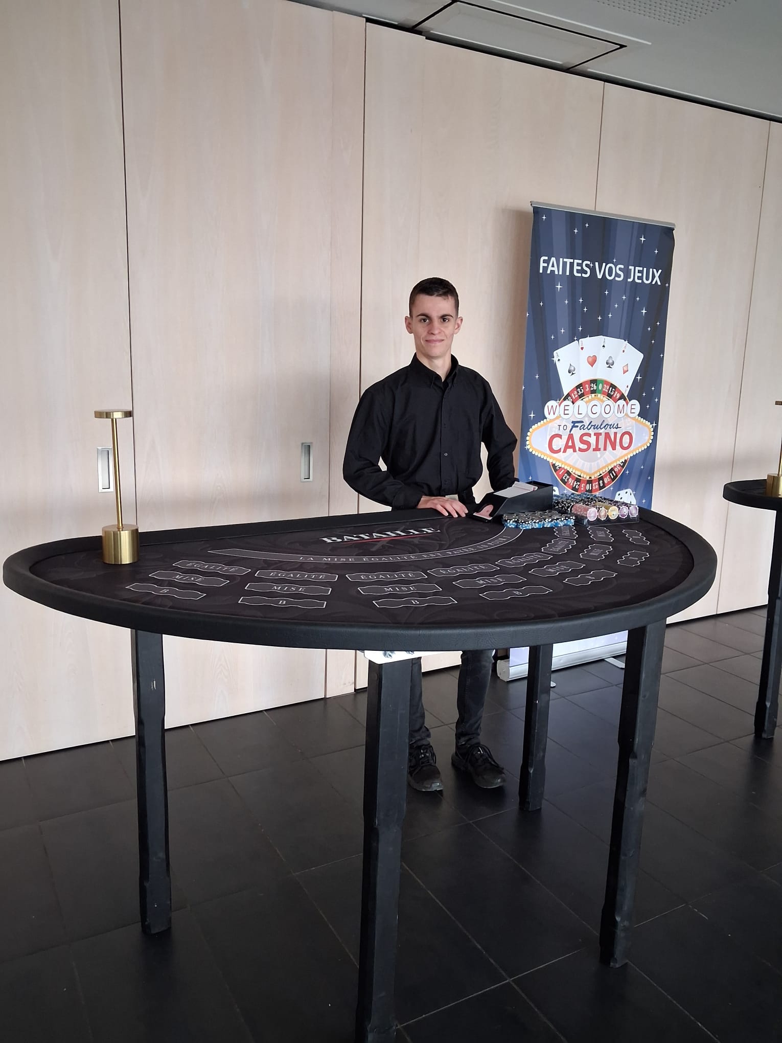 Jeune croupier professionnel souriant derrière sa table de jeu de Bataille lors d'une animation casino d'entreprise à Brest
